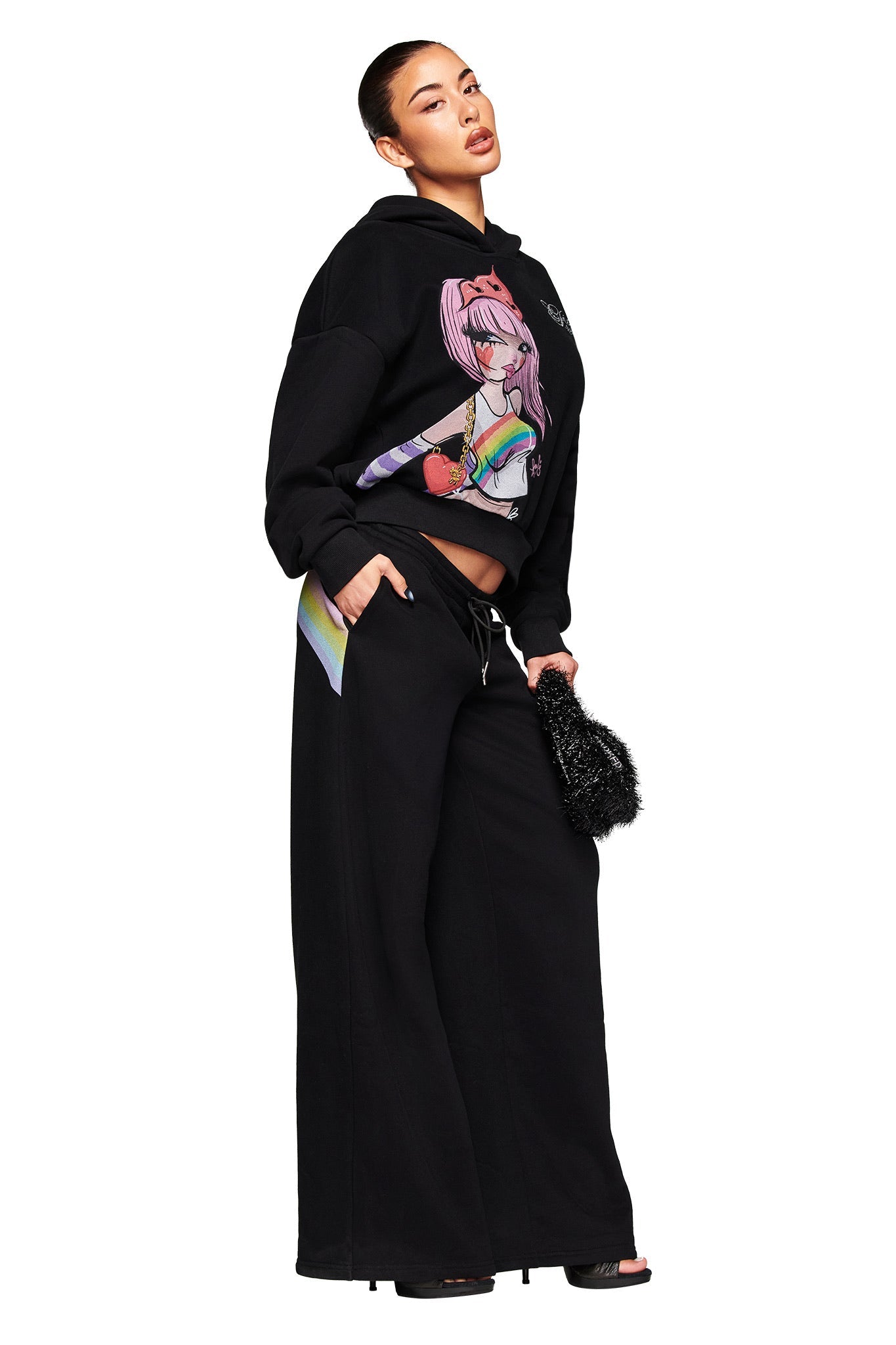 A woman poses confidently in the I.AM.GIA FAFI HOODIE - BLACK, styled with wide-leg black pants, a small black fuzzy bag, and open-toe heels. Her hair is tied back, highlighting the bold, oversized hoodie inspired by Fafinettes.