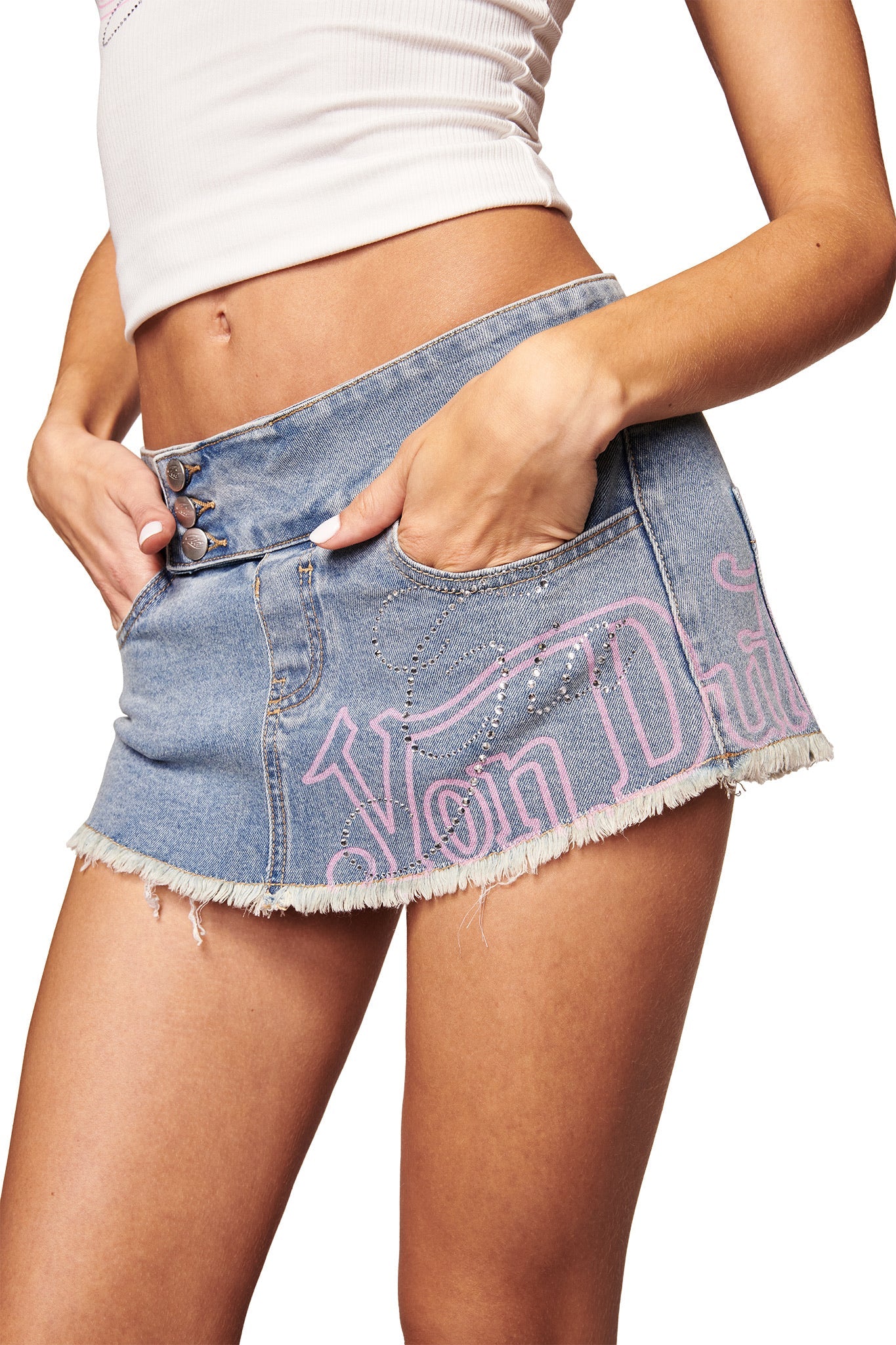 A woman wears the I.AM.GIA X VON DUTCH SYNDRA DENIM MINI SKORT in light wash with pink "Von Dutch" lettering and a white crop top. Her hands are in the skort pockets; the image is cropped from shoulders to mid-thigh.