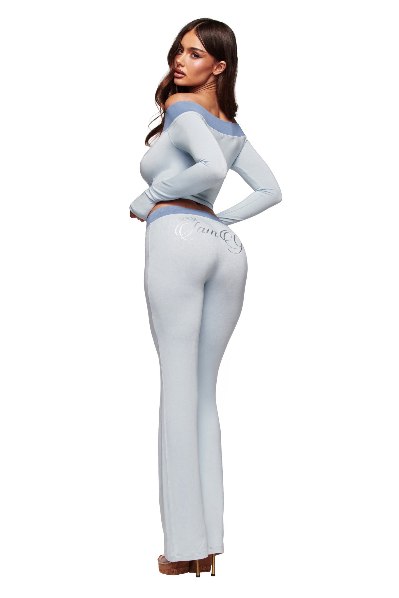 A woman with long brown hair wears I.AM.GIA’s BLARE LOWER RISE TRACKPANT in two-tone powder blue, posed barefoot, turned to show her back and looking over her shoulder against a white backdrop.