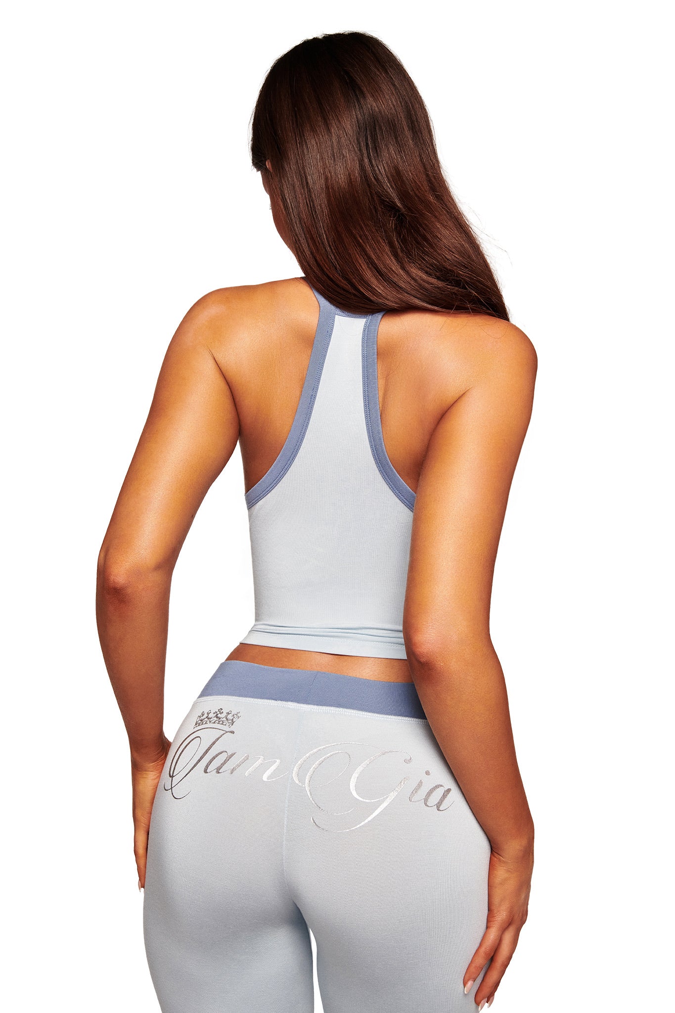 A woman with long brown hair, seen from behind, wears the I.AM.GIA BLARE LOWER RISE TRACKPANT in two-tone powder blue. The sporty pants feature script writing on the back and darker blue trim, set against a white background.