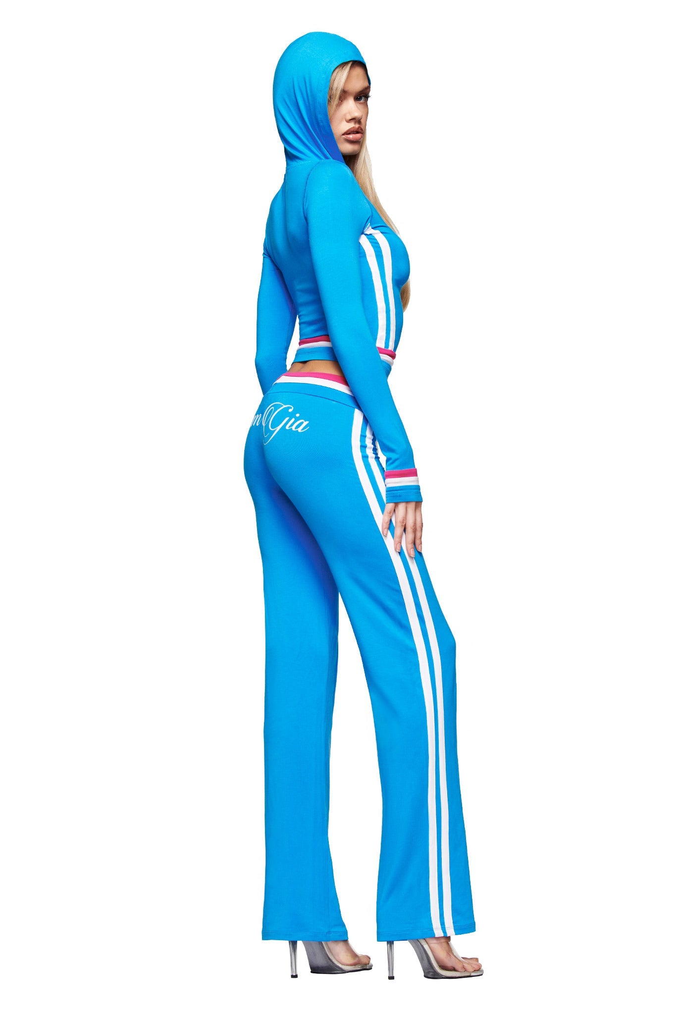 A woman models the I.AM.GIA BLARE STRIPED TRACKPANT in sky blue, featuring straight legs, white side stripes, a low-rise waist, and multicolored trim. She pairs it with silver heels and wears the hood up.
