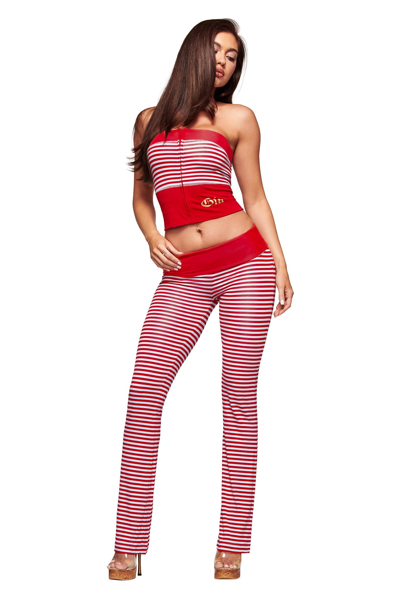 A woman poses in the I.AM.GIA BLARE ZIP BOOB TUBE in red and blue stripe, paired with matching flared pants. She wears tan heels and has long brown hair, set against a white background.