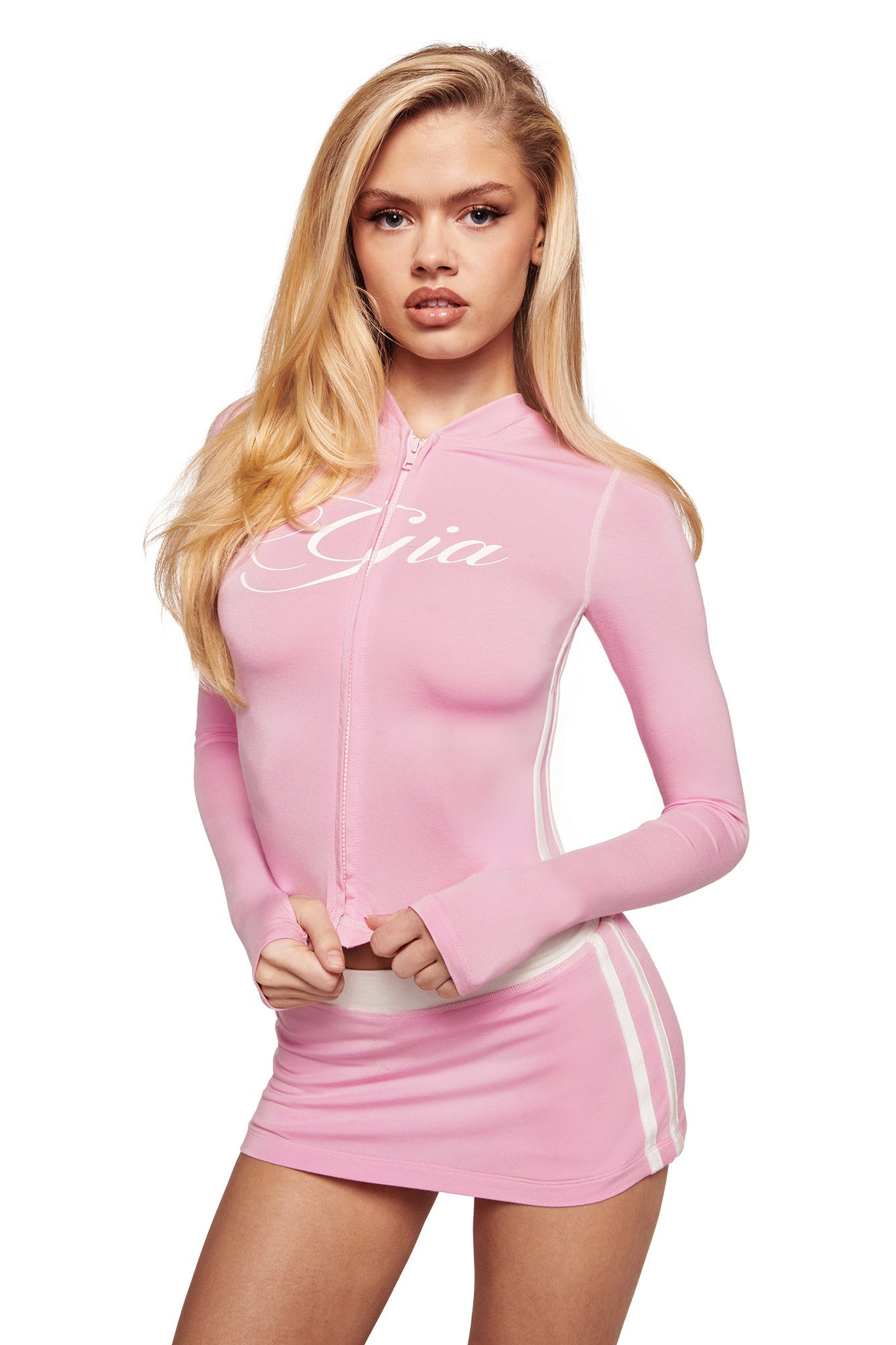 A woman with long blonde hair wears a light pink zip-up hoodie with “Gia” and the I.AM.GIA BLARE PIPING MICRO SKORT in baby pink and white, standing against a plain white background.