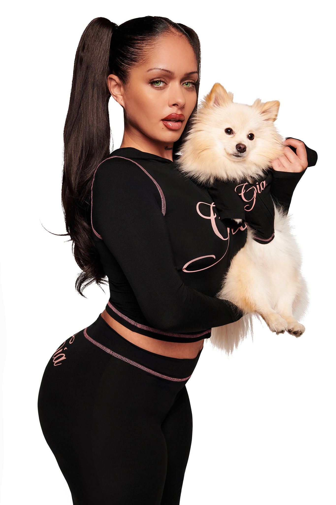A woman with long dark hair in a high ponytail poses against a white background, holding a fluffy white Pomeranian dressed in the I.AM.GIA BLARE BEST FRIEND DOG HOODIE - BLACK. Both wear stylish black matching outfits with pink trim and cursive writing.