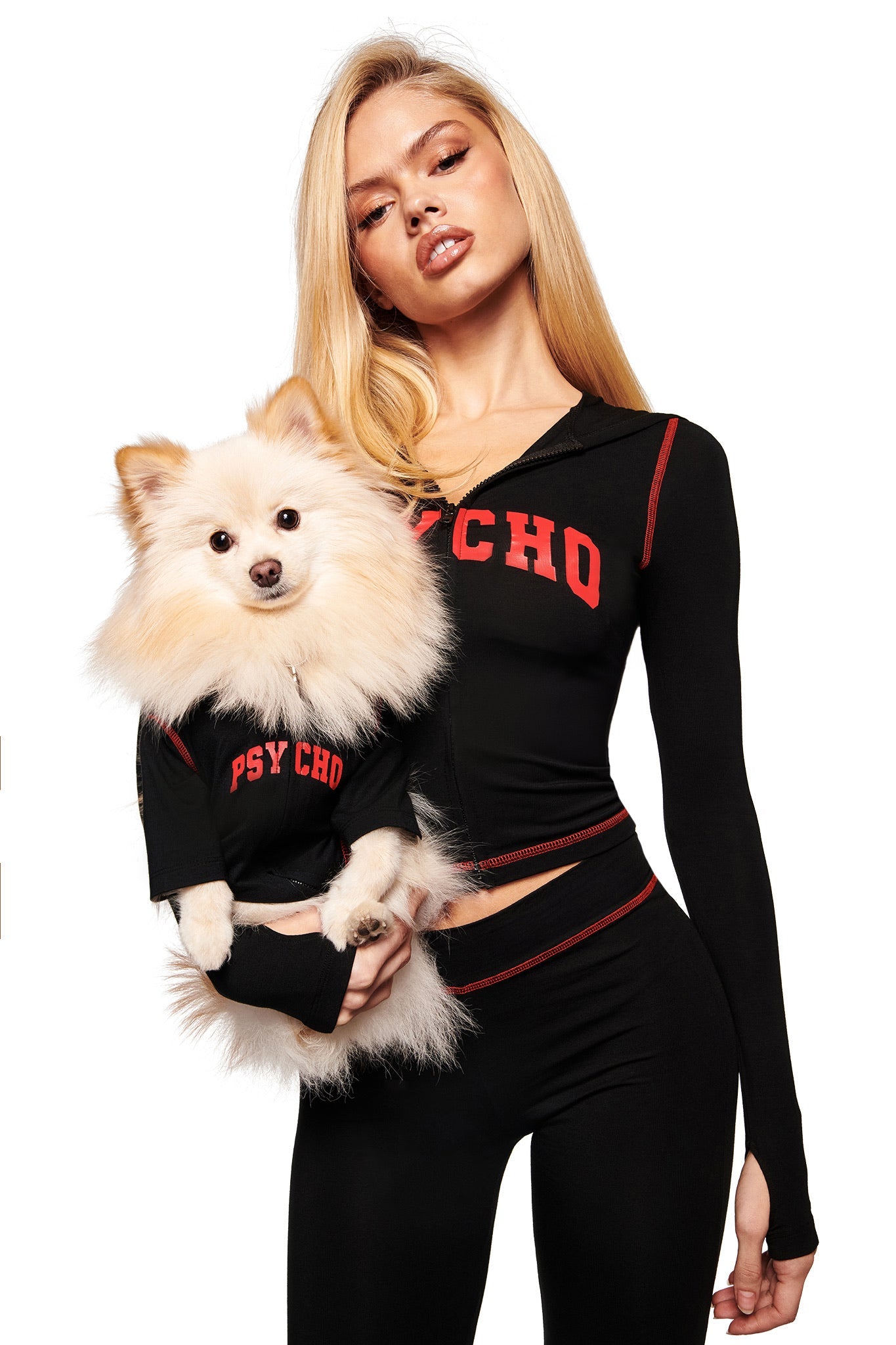 A woman in a black I.AM.GIA outfit with red "PSYCHO" lettering holds a fluffy white dog wearing the BLARE BEST FRIEND DOG HOODIE by I.AM.GIA in black with matching red letters. Both face forward on a plain white background.