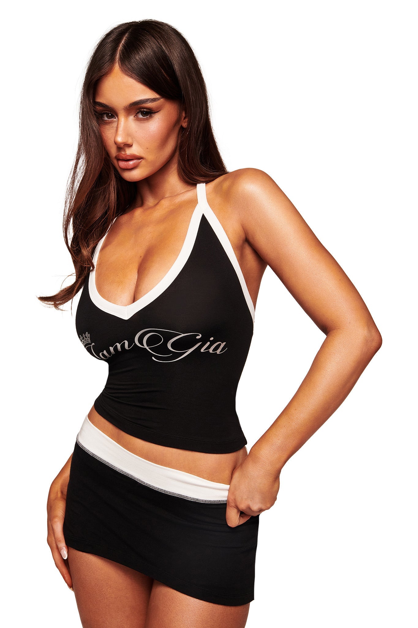 A woman with long brown hair models the I.AM.GIA BLARE MICRO SKORT in black and white, paired with a black halter top featuring white trim and "Tom Gia" lettering, all made from buttery-soft viscose, posing against a white background.