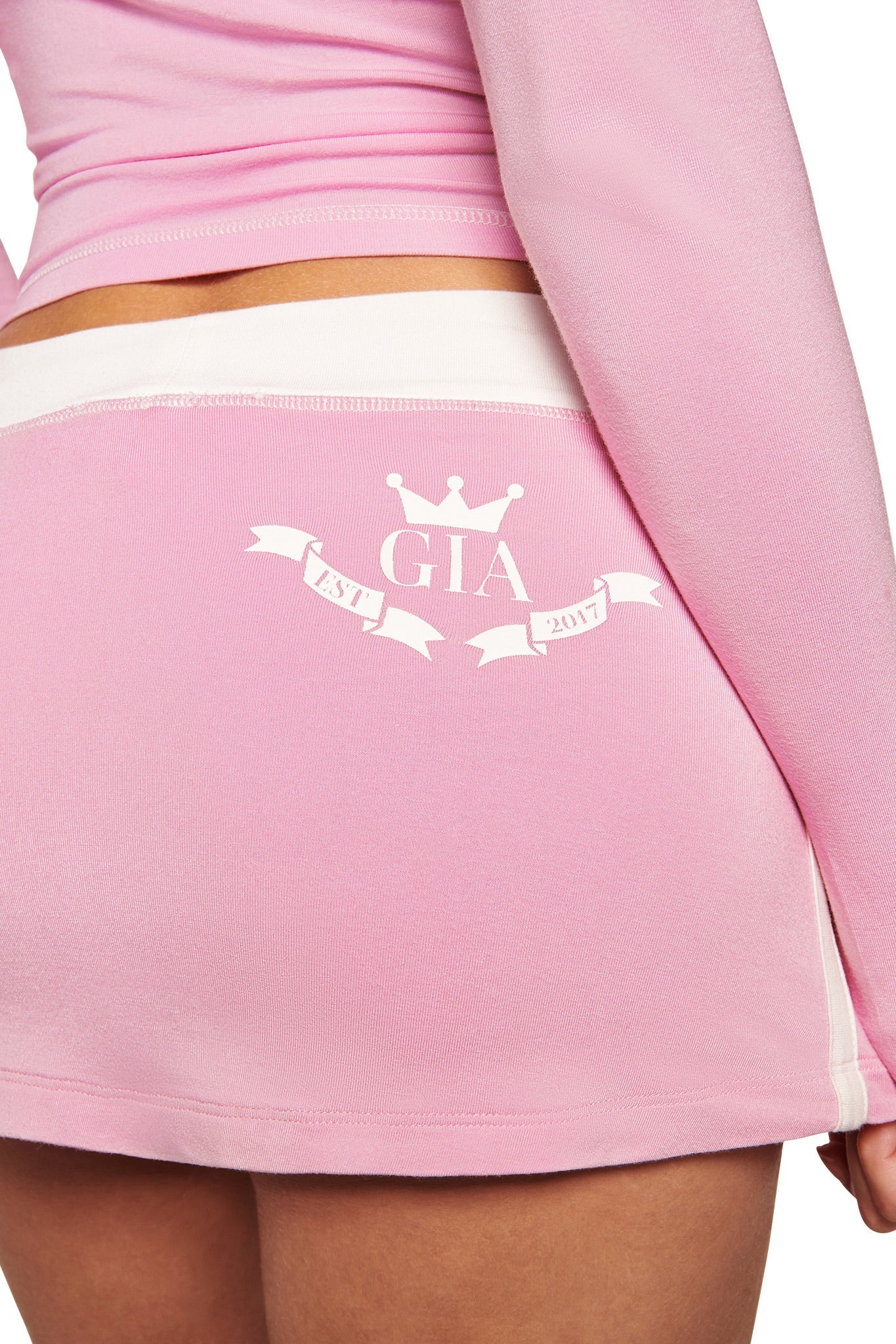 A person wears the I.AM.GIA BLARE PIPING MICRO SKORT in baby pink and white, featuring a white waistband and a back crest with a crown, "GIA," and ribbons reading "Est. 2017," paired with a matching long-sleeve top.