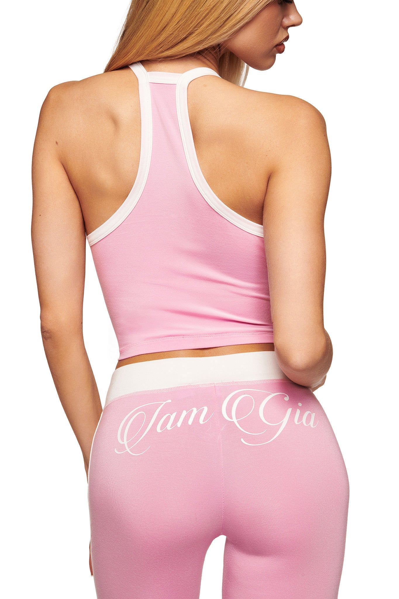 A woman with long blonde hair faces away, wearing the I.AM.GIA BLARE V-NECK TANK in baby pink and white with matching high-waisted pants featuring “I.AM.GIA” script on the back, set against a white background.