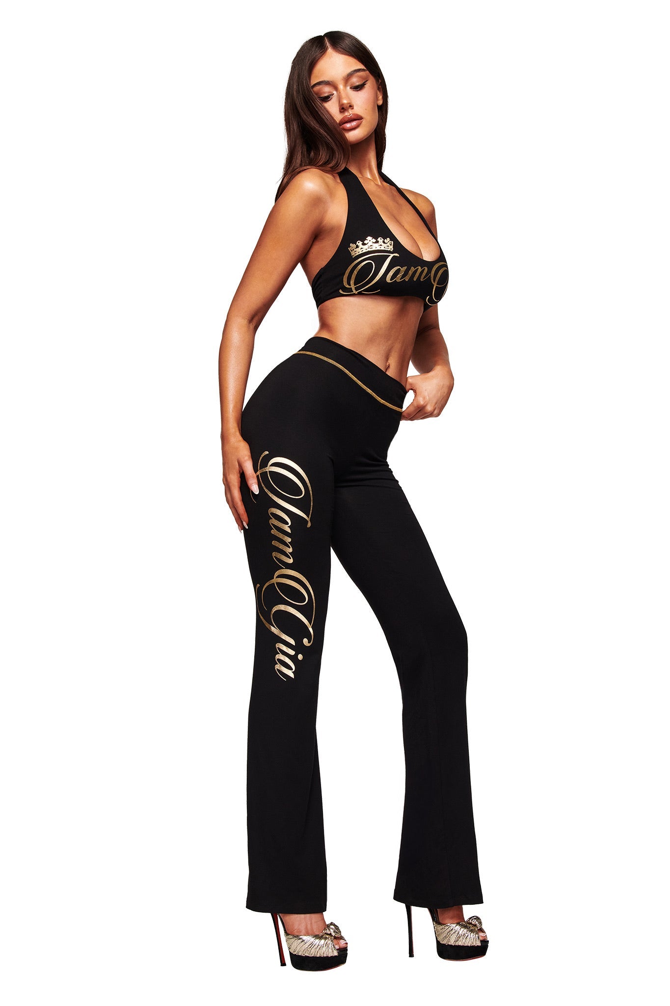 A woman models the I.AM.GIA BLARE FORM TRACKPANT in black and gold with a matching branded top, high heels, and poses against a white background.