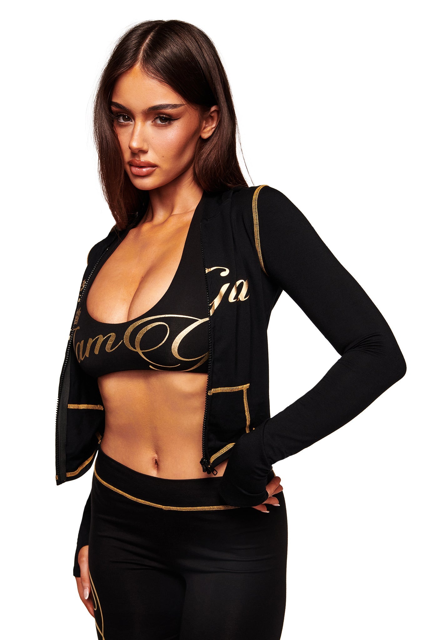 A woman wears the I.AM.GIA BLARE FORM HALTER CROP TOP in black and gold, paired with matching leggings, standing against a white background.