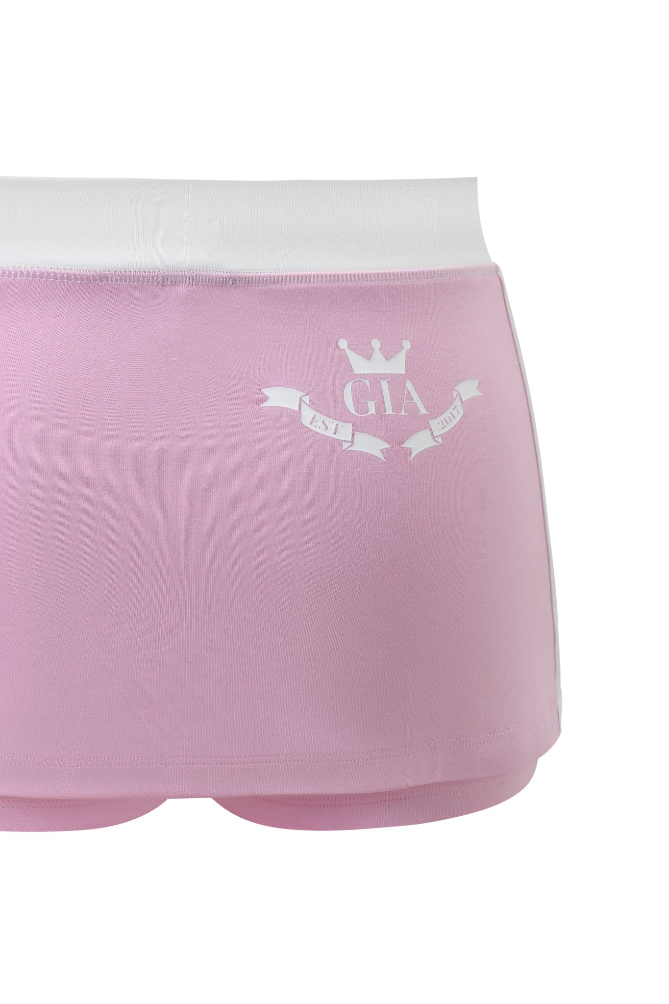 A close-up of the I.AM.GIA BLARE PIPING MICRO SKORT in baby pink and white, featuring a white waistband, crown graphic, "GIA" text, and a ribbon design with "EST." and "2021" printed on it.