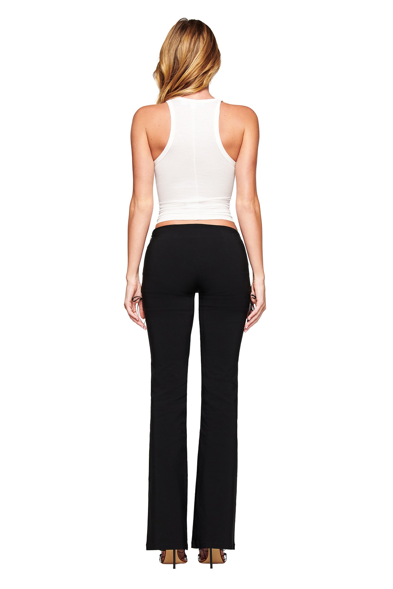 A woman faces away from the camera in a white PENELOPE TANK - PSYCHO by I.AM.GIA and black flared pants with high heels, standing against a plain white background.