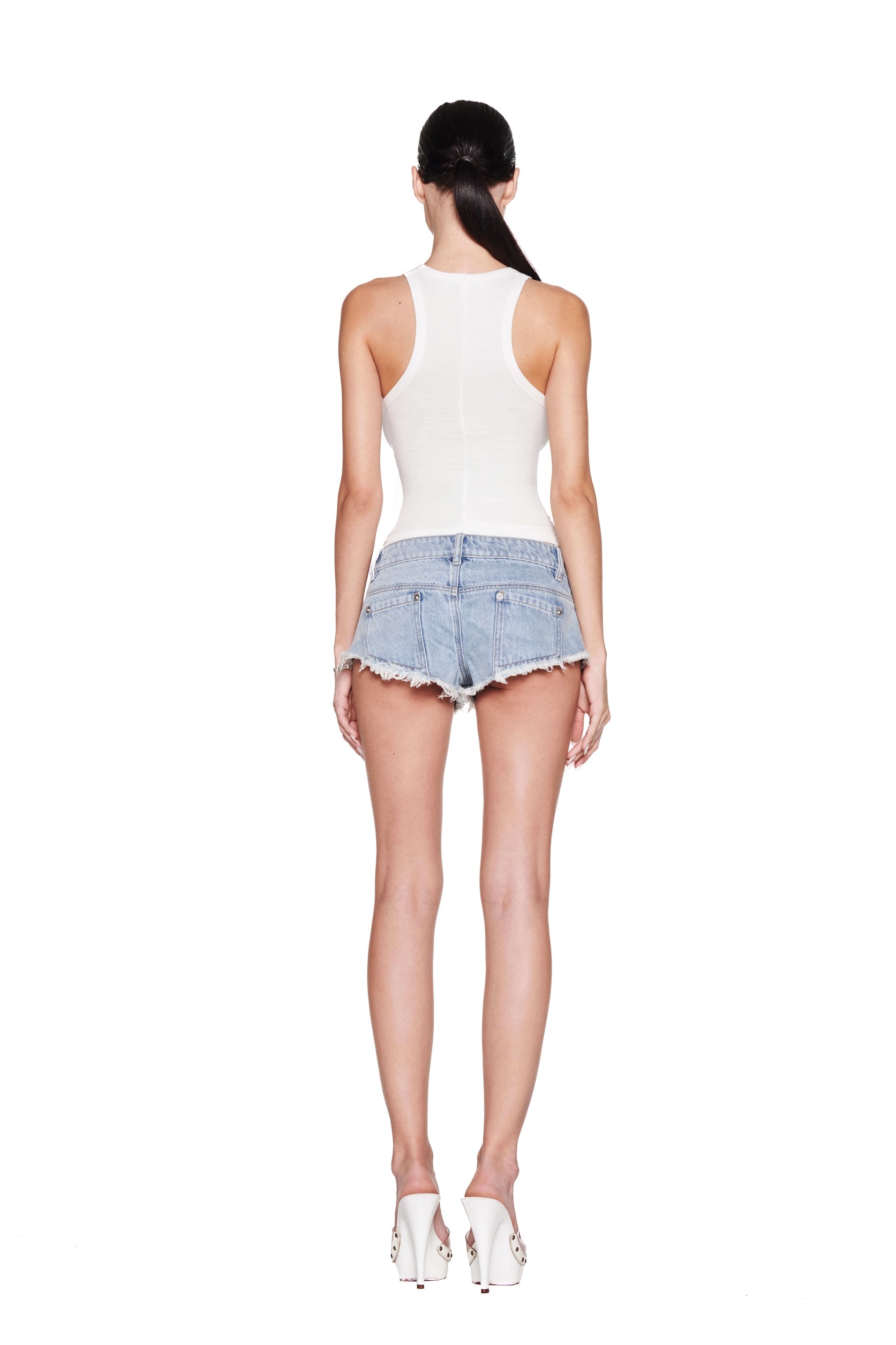 A person with long hair in a ponytail wears a PENELOPE TANK - GIA GIRL from I.AM.GIA, frayed denim shorts, and white high-heeled mules, standing against a white background.