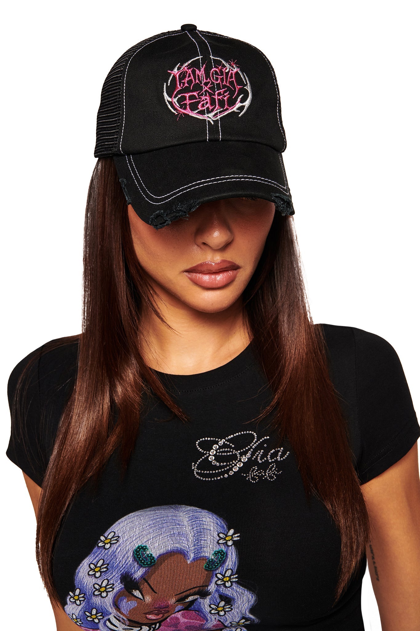 A woman with long brown hair looks down, wearing the I.AM.GIA FAFI HAT - BLACK—a distressed black trucker hat with an embroidered logo—and a black tee featuring a cartoon deer with flowers.