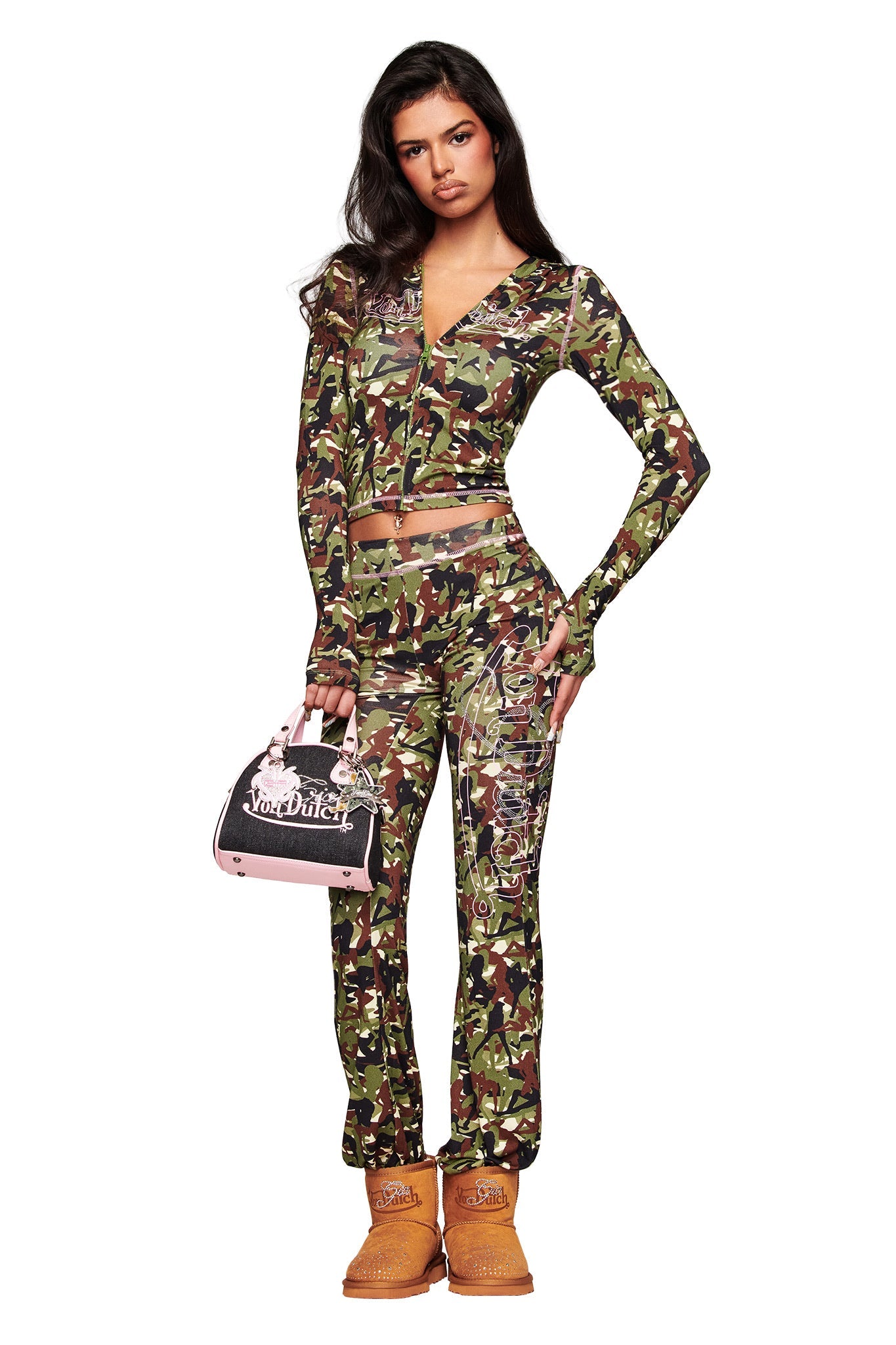 A woman with long dark hair and a neutral expression wears the I.AM.GIA X VON DUTCH BLARE HOODIE in Gia Girl Camo, matching pants, brown boots, and holds a black and pink Von Dutch handbag.