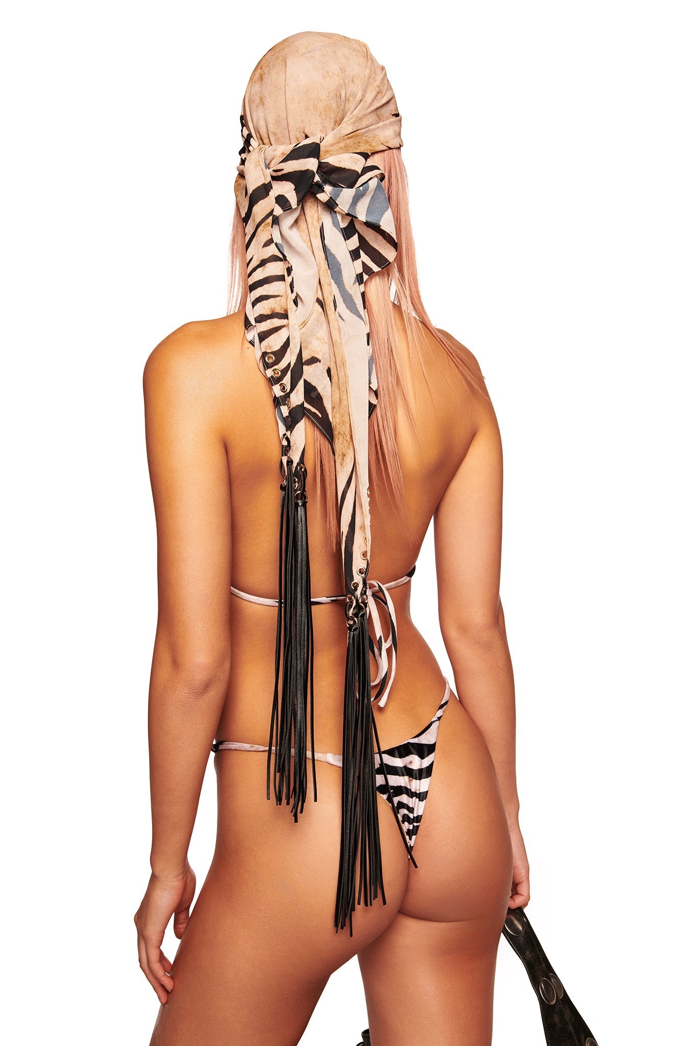 A person with long light hair is seen from behind, wearing a zebra-print bikini and the I.AM.GIA ALLUNA MULTI-WEAR TASSEL SCARF - WILD SIDE, featuring animal print with fringe details and tassels down the back.