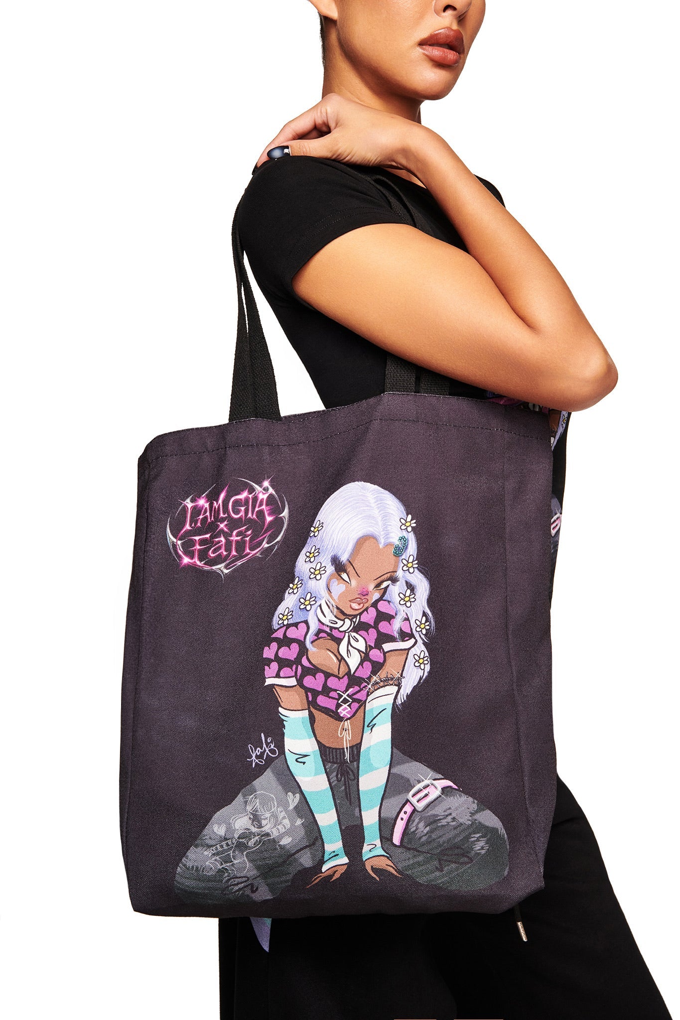 A woman in a black shirt carries the FAFI TOTE BAG - BLACK by I.AM.GIA, featuring exclusive colorful artwork from the FAFI collaboration with a doll-like character, striped stockings, and “TAMGIA FATI” text.