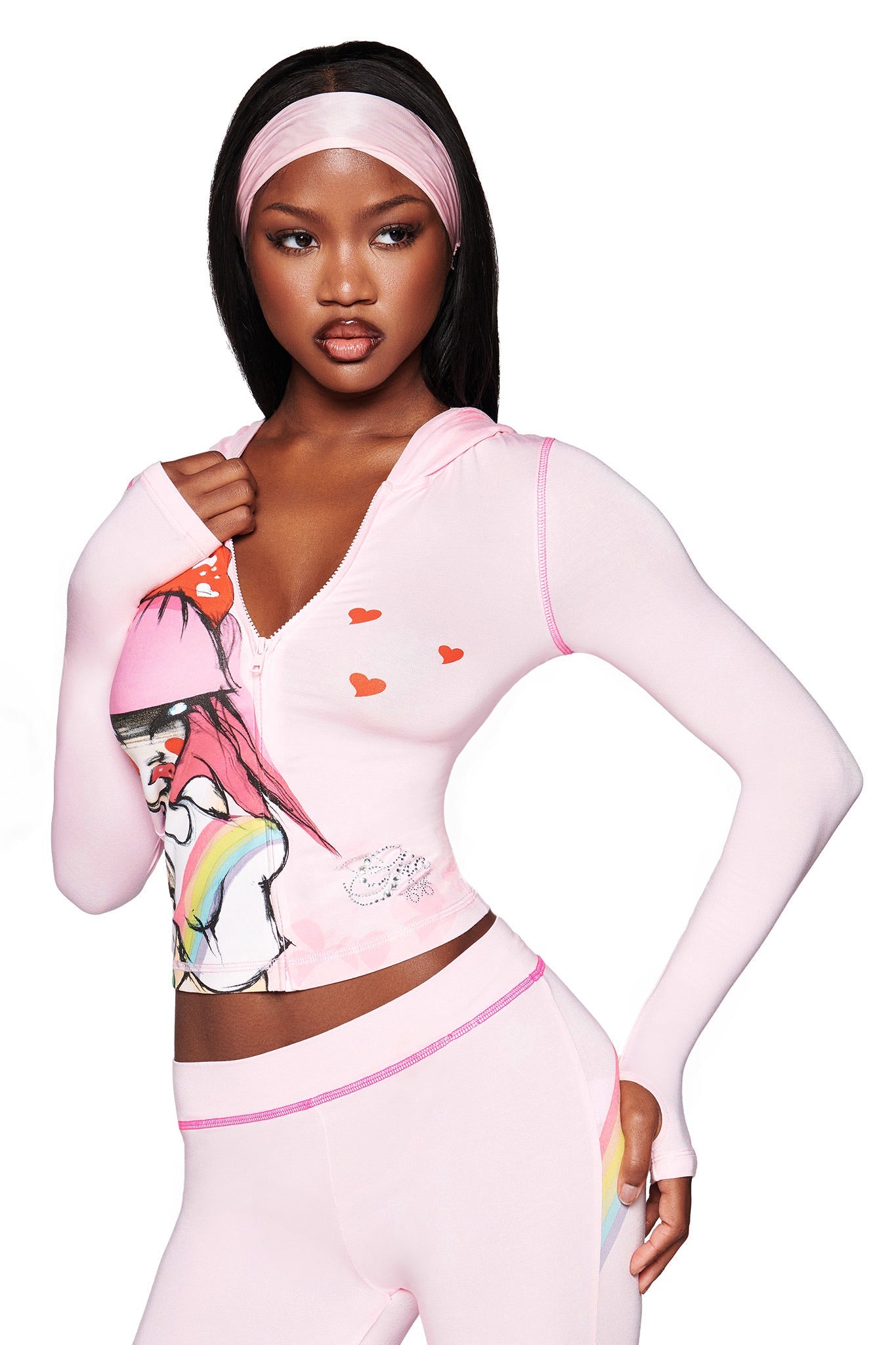 A woman models the I.AM.GIA FAFI BLARE HOODIE in baby pink, featuring illustrated graphics, hearts, and diamanté details, paired with matching leggings and a pink headband against a white background.