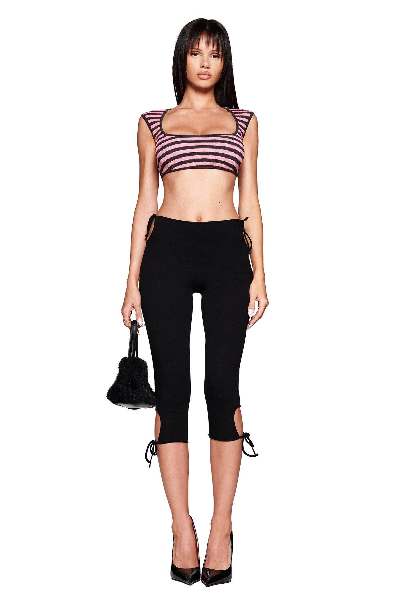 A woman poses against a white background wearing I.AM.GIA's YUNA CAPRI - BLACK mid-rise, body-con capri pants with side ties, paired with a pink and black striped crop top, black heels, and holding a small black bag.