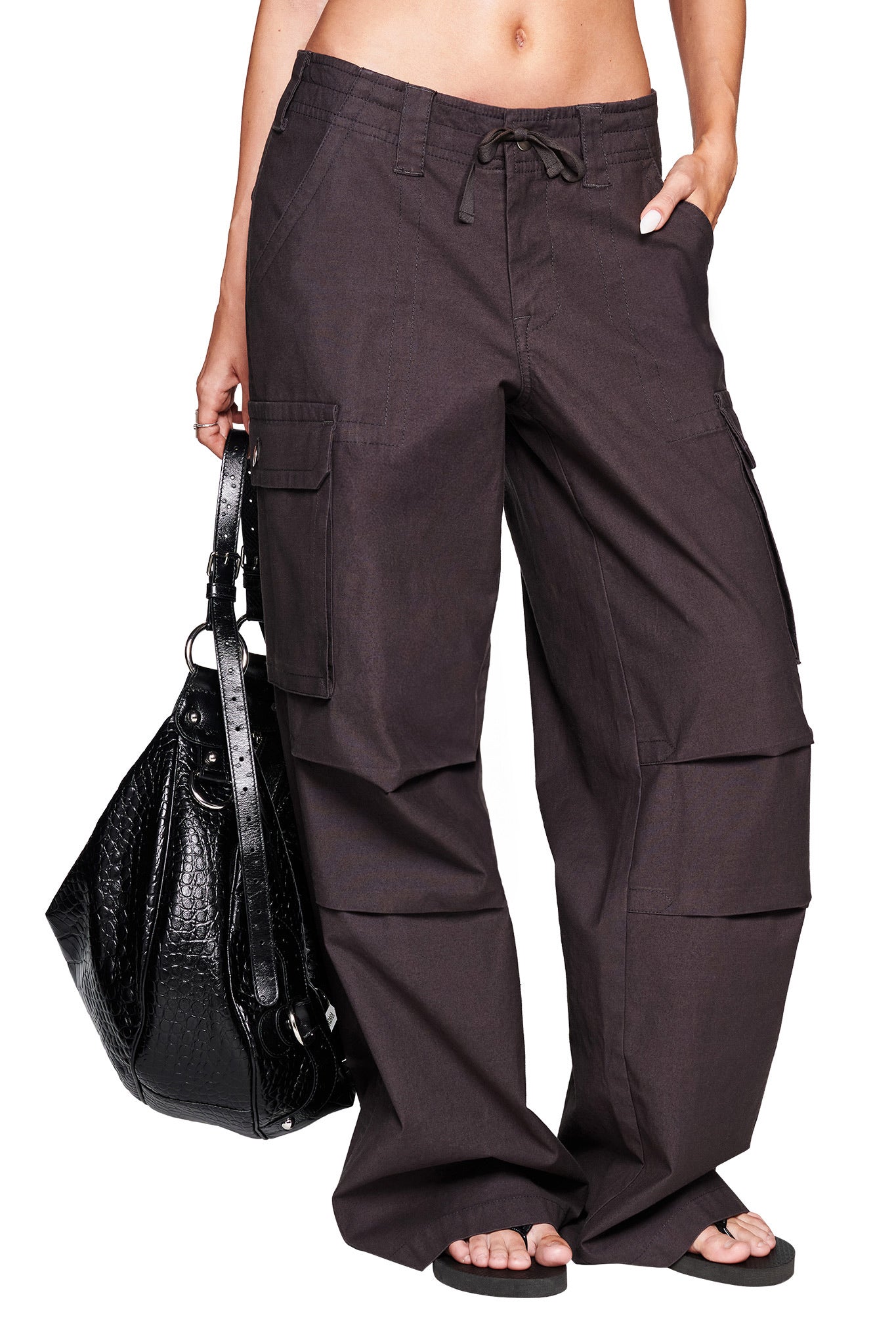 A person wearing the I.AM.GIA JAXXON PANT in grey with a baggy leg silhouette and flip-flops holds a large black textured handbag, with only their lower torso and legs visible.