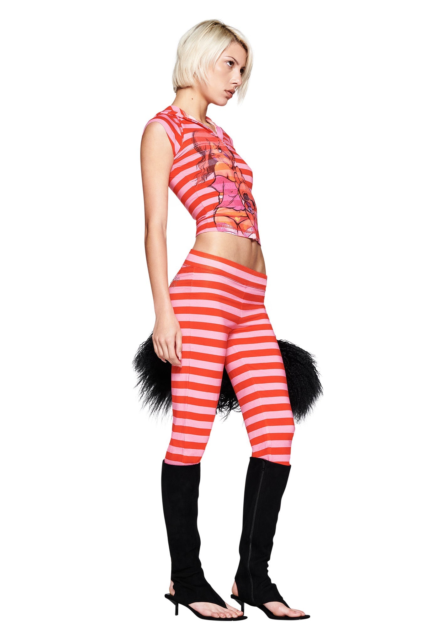 A woman with short blonde hair stands in profile against a white background, wearing the I.AM.GIA FAFI BLARE CAP SLEEVE HOODIE in pink stripe, paired with matching leggings and black high-heeled open-toe boots.