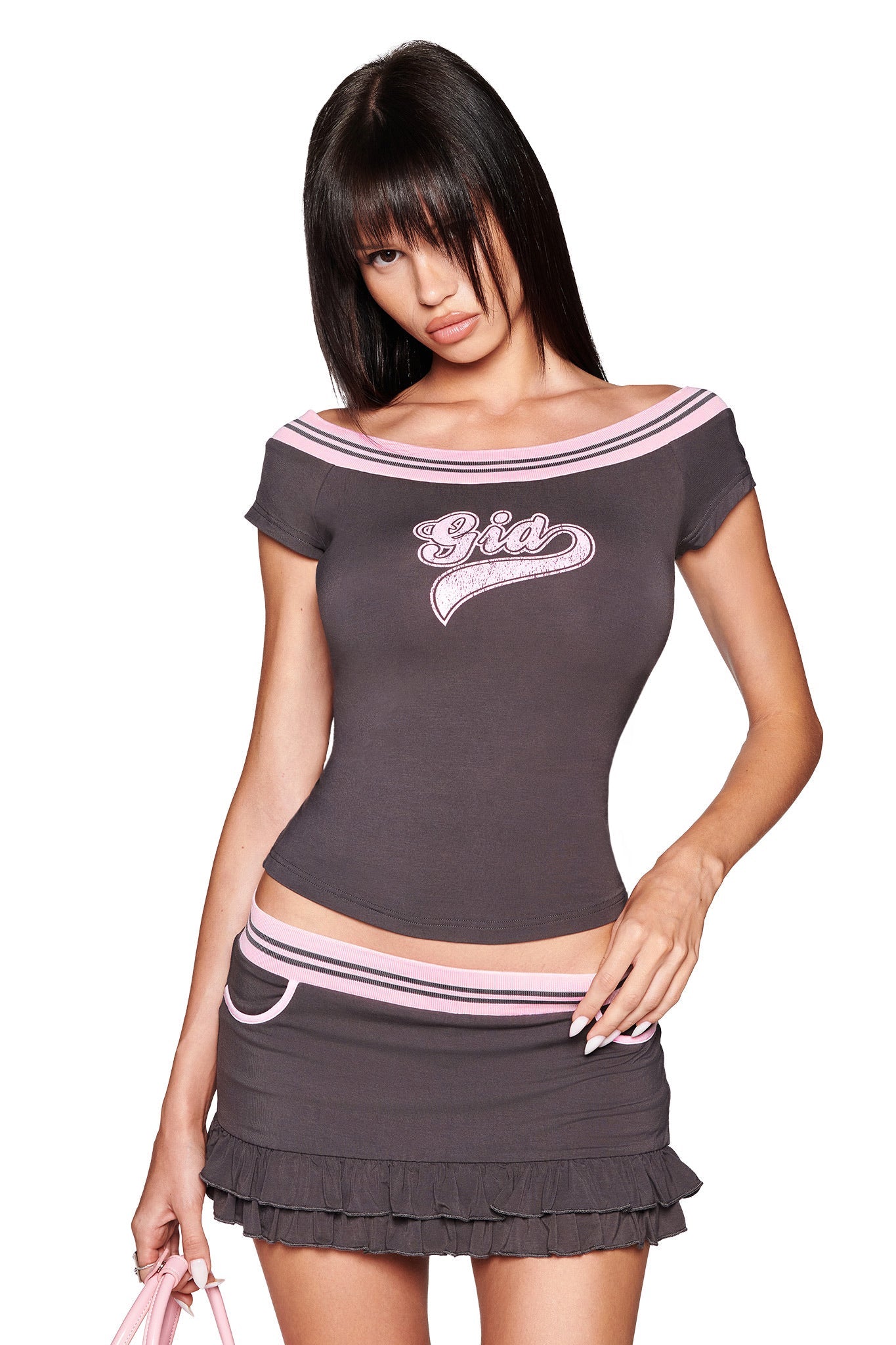 A woman with straight dark hair wears the I.AM.GIA ELLIS SKORT in grey, paired with a matching off-shoulder charcoal top with pink trim and "Gia" on it. She holds a light pink item in her left hand.