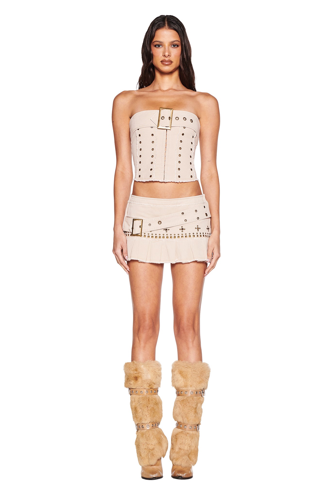 A woman faces forward against a white background, wearing the I.AM.GIA MASON MINI SKIRT in tan with a low rise and pleats, paired with a beige strapless top featuring buckles and grommets, and fluffy tan boots with buckles.