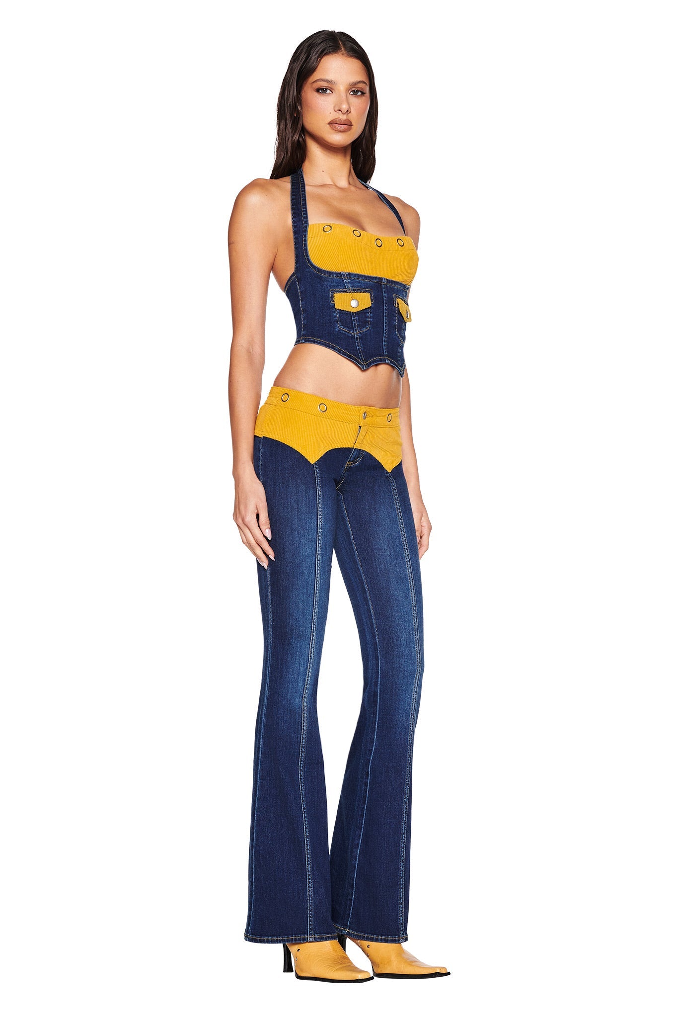 A woman wears the I.AM.GIA TOMMIE JEAN - DARK WASH: a strapless denim crop top and flared pants with a corduroy waistband, paired with yellow heeled boots, all set against a plain white background.