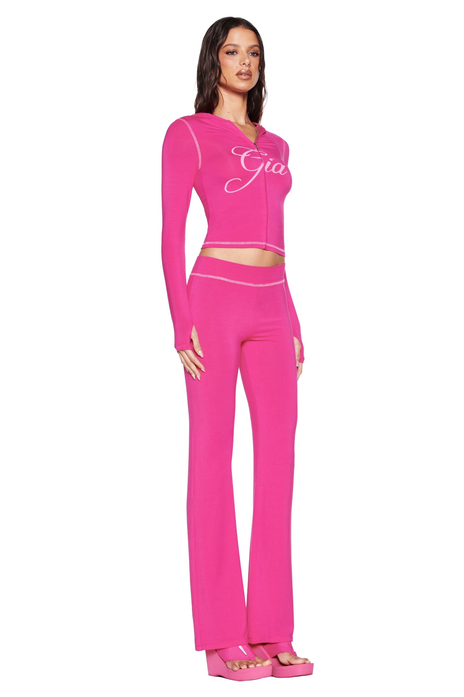 A woman in a vibrant hot pink outfit stands against a white backdrop wearing a "Gia" long-sleeve top, matching I.AM.GIA Blare Trackpant in Hot Pink, and platform sandals.