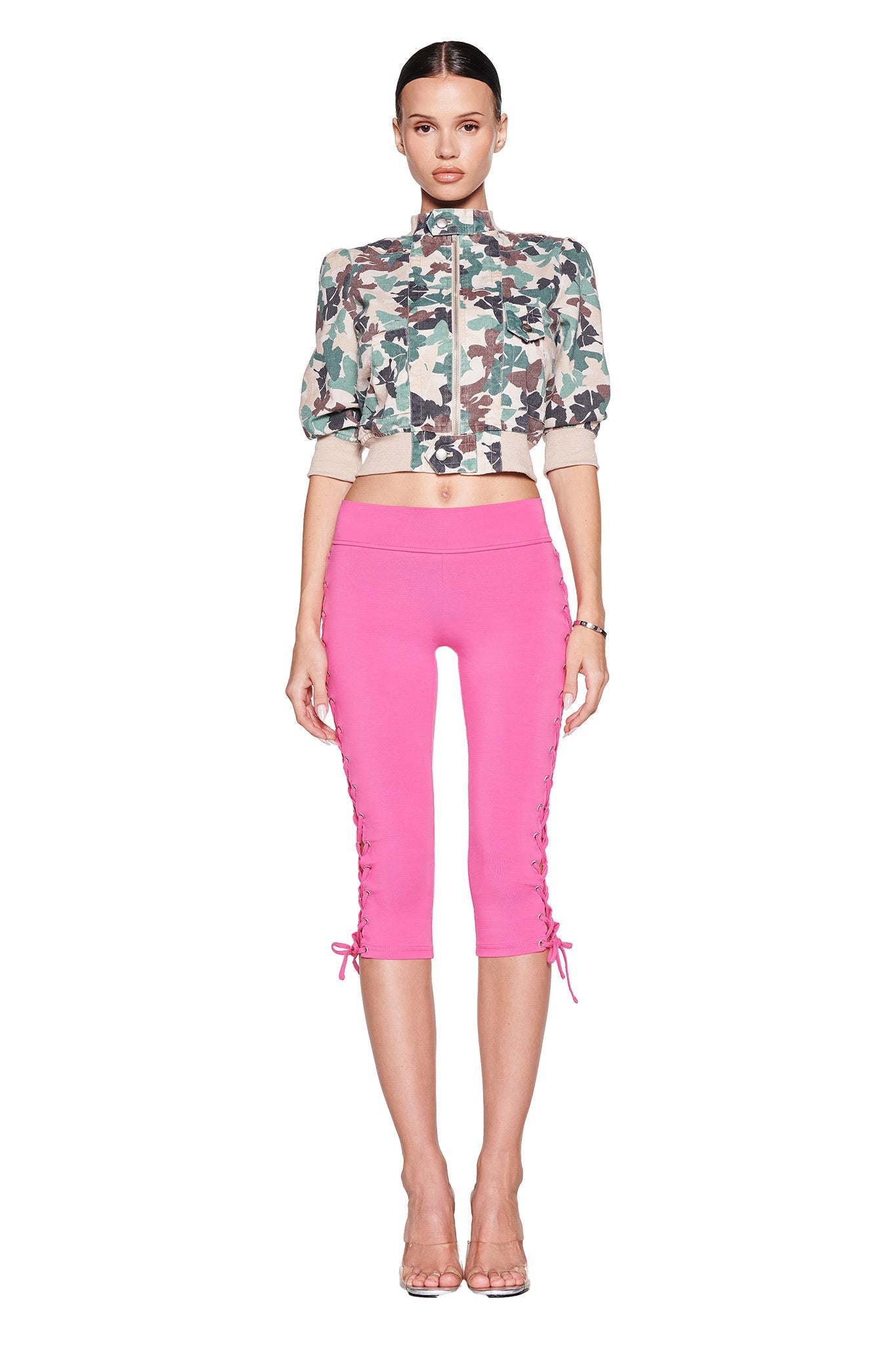 Against a white background, a person dons the I.AM.GIA camo-patterned cropped jacket and TILA CAPRI - HOT PINK jersey capri pants with lace-up details. Their slicked-back hair and clear high heels complete the chic, confident look.