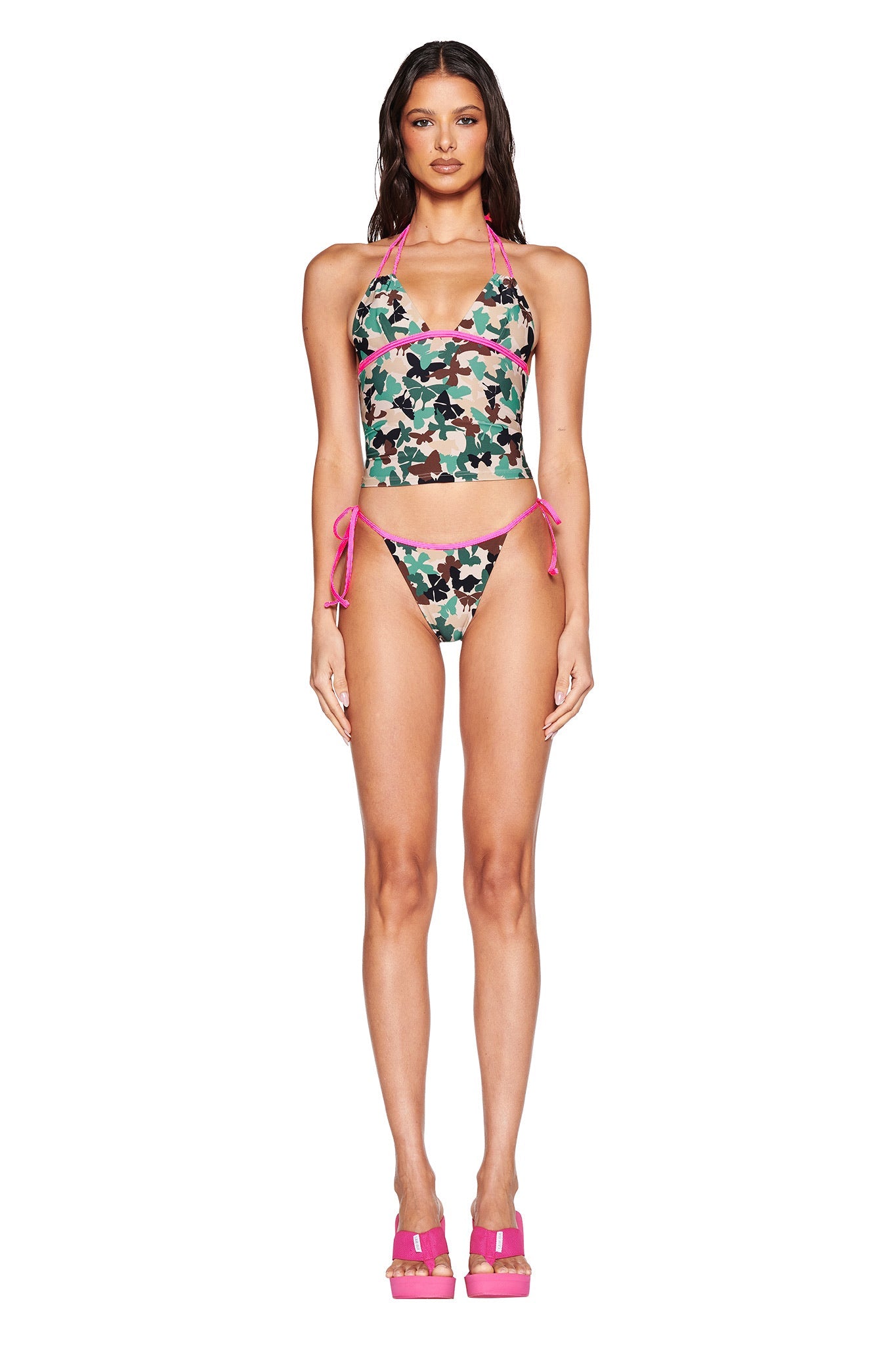 A woman confidently models the SEDONA BIKINI BOTTOM - FLUTTER CAMO by I.AM.GIA, featuring cheeky, low-rise design with pink strap accents. She stands in pink platform heels against a white background, exuding effortless style.
