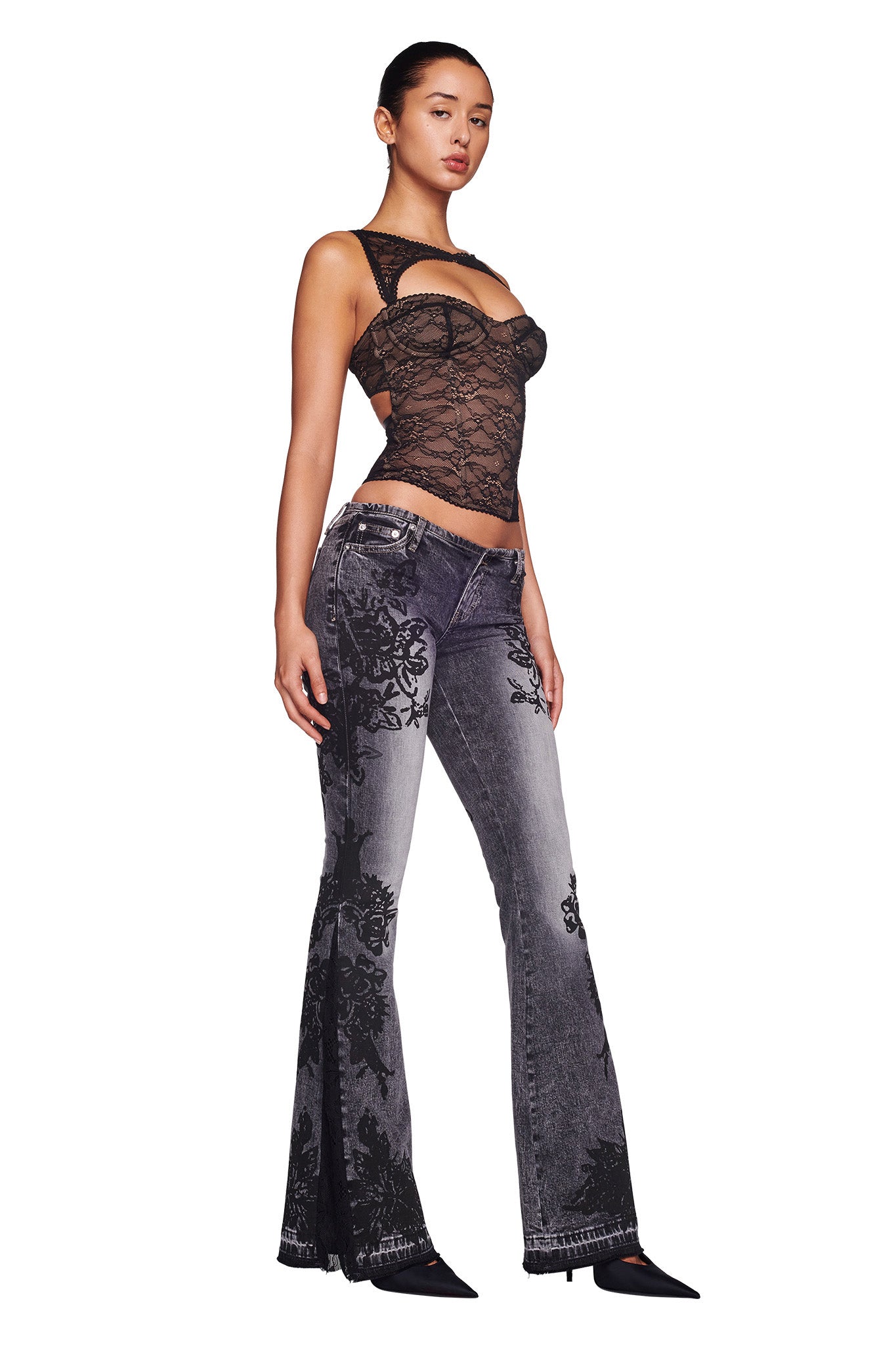 A woman stands against a white background, exuding confidence in her I.AM.GIA KIANA BUSTIER - BLACK and gray jeans with black floral patterns. She completes her outfit with black pointed-toe heels. Her hair is pulled back as she boldly looks into the camera.