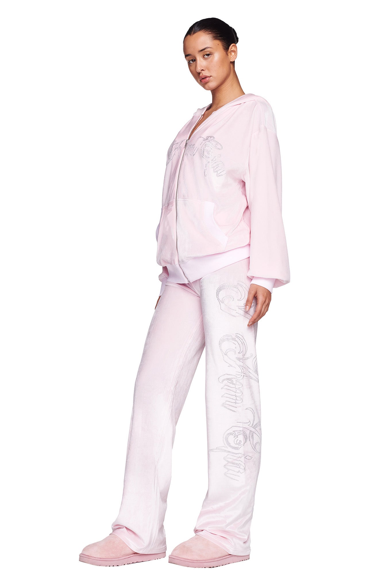 A person stands wearing a matching I.AM.GIA tracksuit in icy pink. The outfit includes the DARIA PANT - PINK: ICY PINK and a hooded jacket with a zipper, paired with pink slippers. With their hair tied back, the person gazes confidently at the camera, showcasing the loose-fitting pants adorned with decorative designs.
