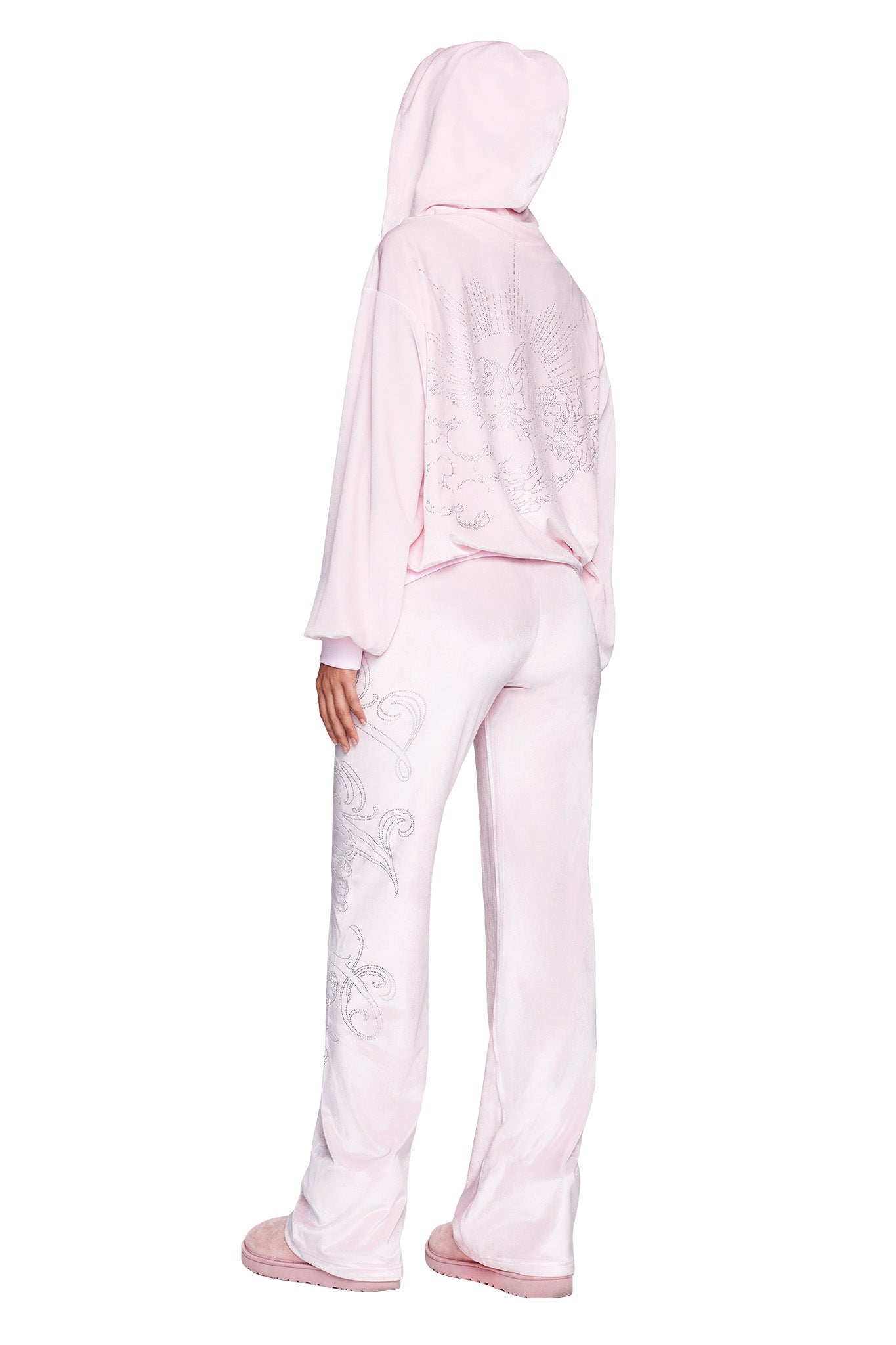 Person wearing an I.AM.GIA DARIA PANT in Icy Pink with a matching velour hoodie, viewed from the back. The tracksuit showcases intricate embroidery on the back of the hoodie and along the sides of the pants. The person completes their look with matching pink slippers.