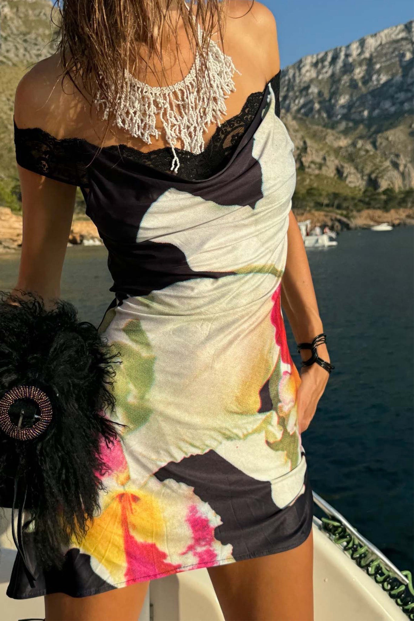 A woman wears the I.AM.GIA THEODORA MINI DRESS - BLACK and a beaded necklace, standing on a boat with a black clutch. Mountains and water are in the sunny background.