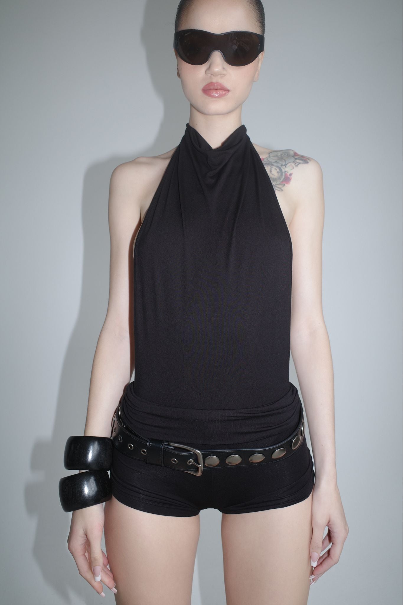 A person wearing sunglasses and the I.AM.GIA KAIRA PLAYSUIT - BLACK stands against a plain light gray background, accessorized with a studded belt and chunky black bracelets.