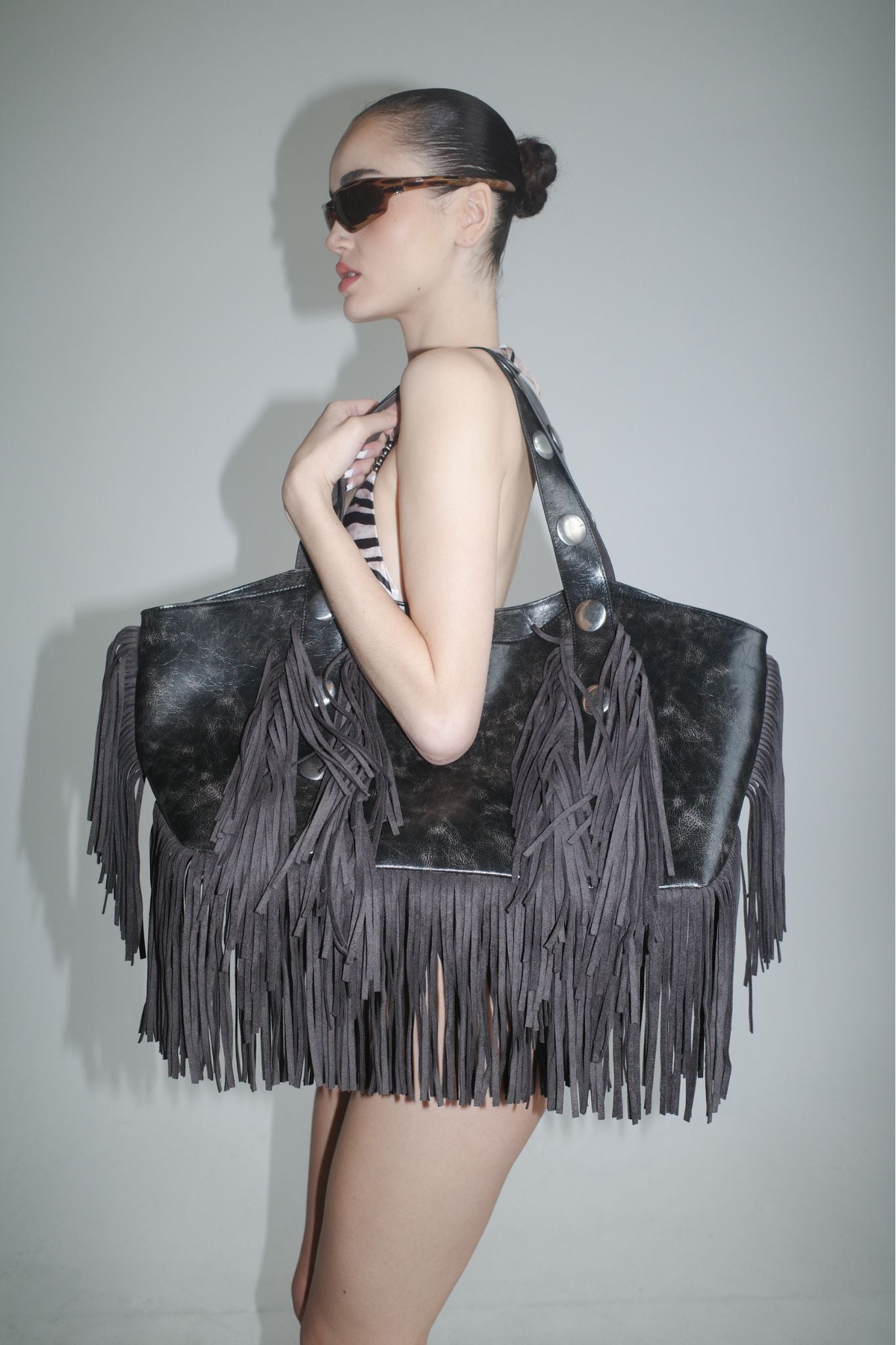 A woman in sunglasses and a striped swimsuit poses in profile, holding the I.AM.GIA CHROMA FRINGE BAG - BLACK with fringe detail against a plain gray background.