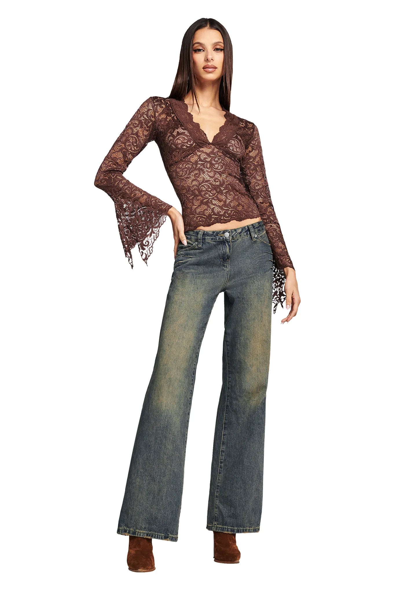 A woman stands confidently against a white background, wearing the GEMMA TOP - NEUTRAL : CHOCOLATE by I.AM.GIA, a long-sleeved, lacy, brown V-neck top with flared cuffs paired with faded blue jeans. She completes the look with brown heeled shoes. Her long, straight hair frames her face as she poses with one hand on her hip.
