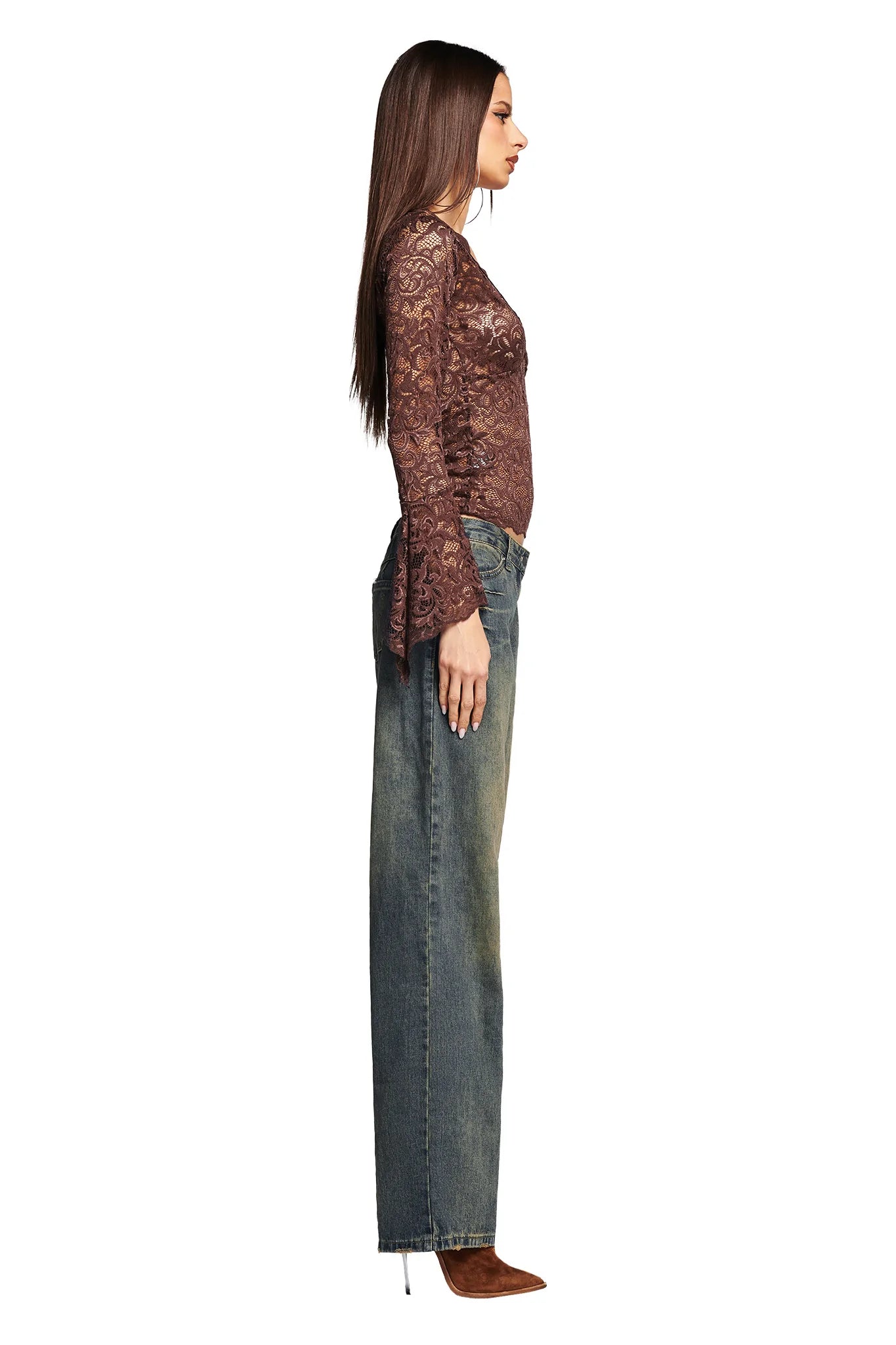 A woman with long brown hair is standing in profile against a white background, showcasing her chic I.AM.GIA ensemble. She is wearing the GEMMA TOP - NEUTRAL : CHOCOLATE, a long-sleeve brown lace top, paired with blue wide-leg jeans and brown pointed-toe heels. Her hands are down by her sides.