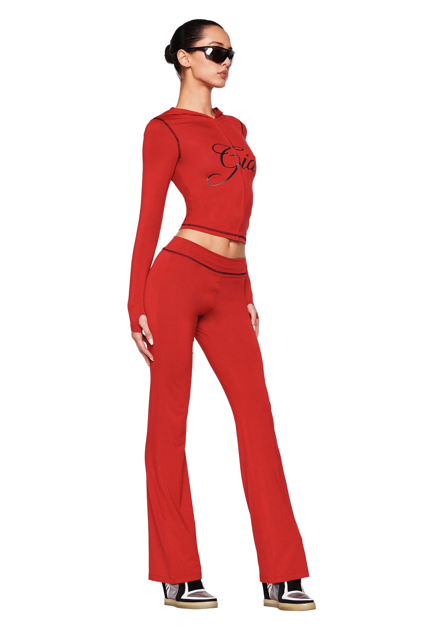 A person in a sleek, all-red outfit stands confidently, wearing a long-sleeve crop top with "I.AM.GIA" emblazoned on it and matching BLARE TRACKPANT - RED. They have dark sunglasses and their hair is pulled back as they pose sideways for the camera.