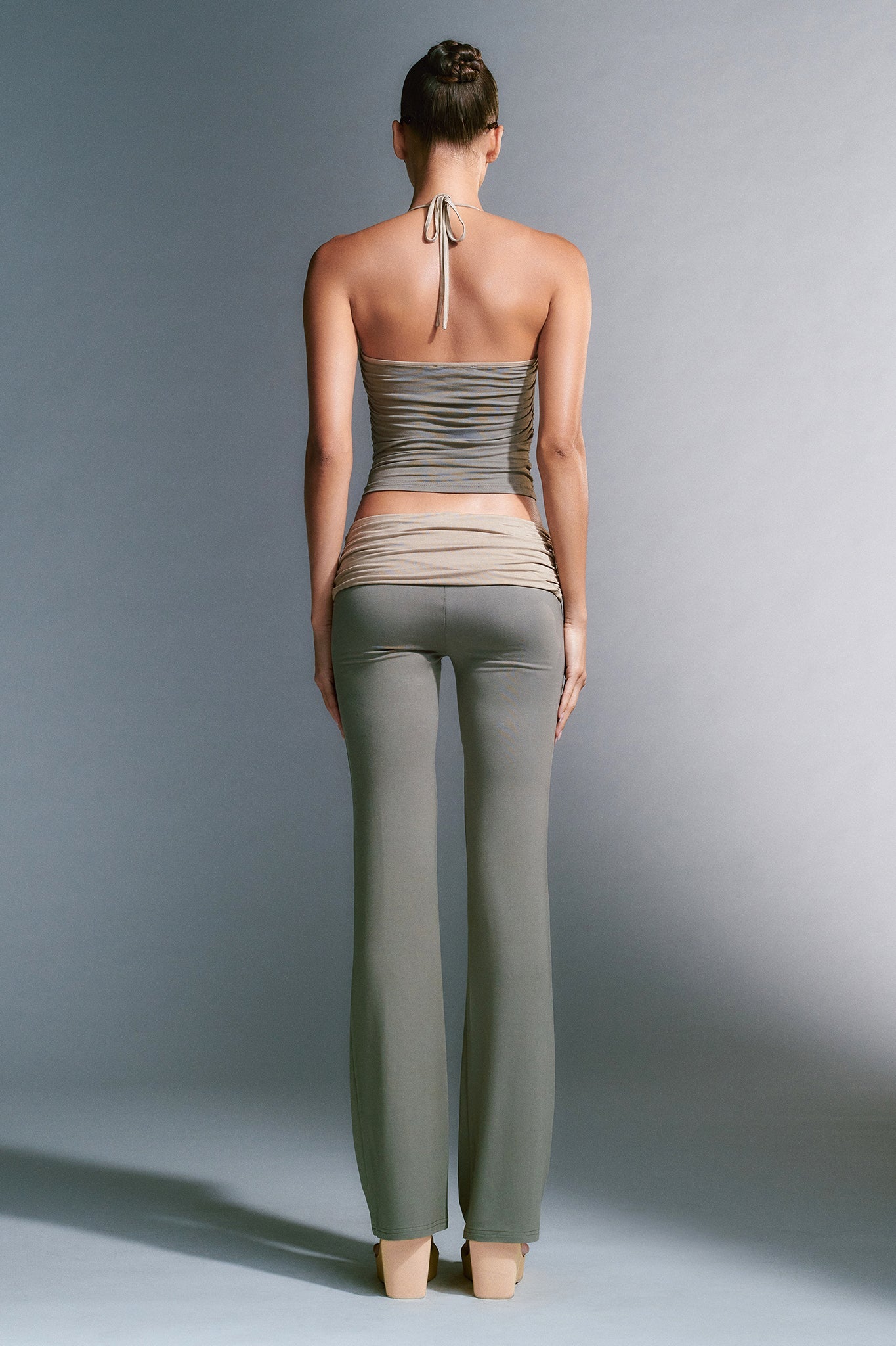 A woman stands with her back to the camera, wearing a backless, halter neck, striped top and the tight-fitting JANNIS PANT - GREEN by I.AM.GIA. Her hair is neatly tied up in a bun. The background is plain and gray with subtle shadows.