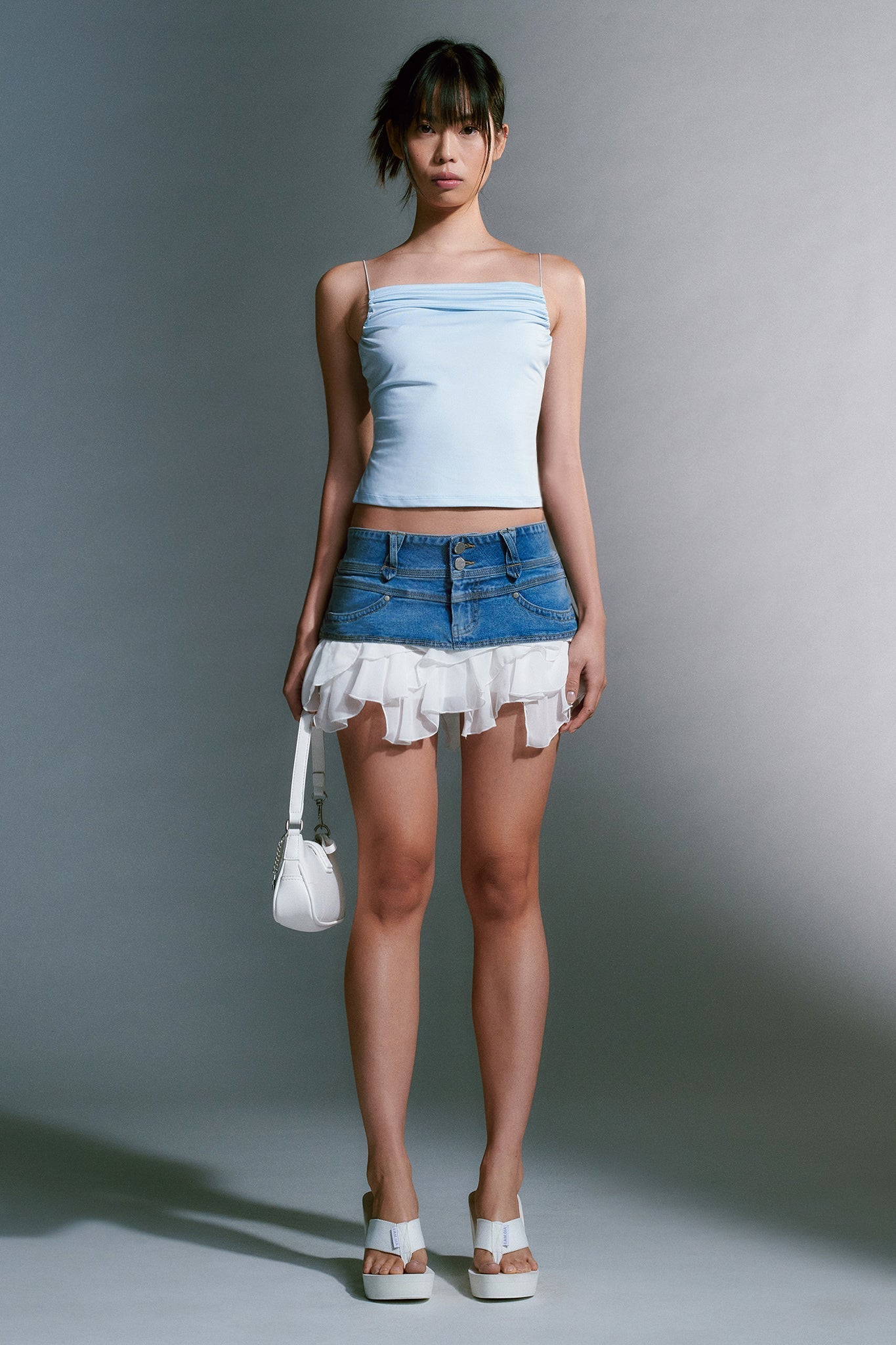 A woman stands against a neutral background, wearing an I.AM.GIA light blue spaghetti strap top and a short denim skirt with a white ruffle hem. She holds a small white handbag and completes her look with the SHELBY FLIP FLOP in white from I.AM.GIA. Her hair is styled in a casual updo.