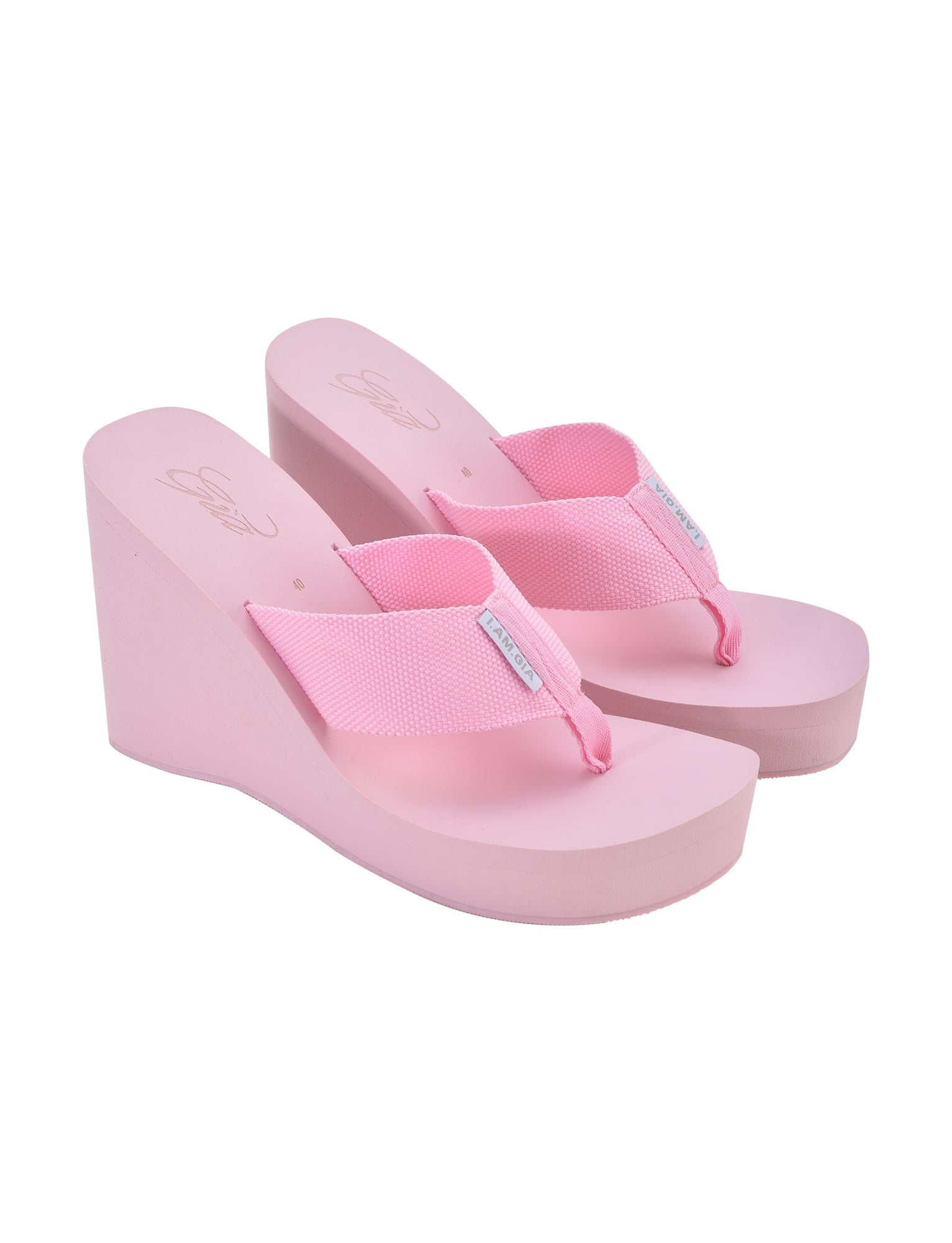 A pair of pink wedge sandals with fabric toe thongs. The wedge heels are solid pink and the toe straps are made of textured fabric, also in pink, featuring a small white tag on each strap stamped with "I.AM.GIA." The brand name "I.AM.GIA" is imprinted on the insole.